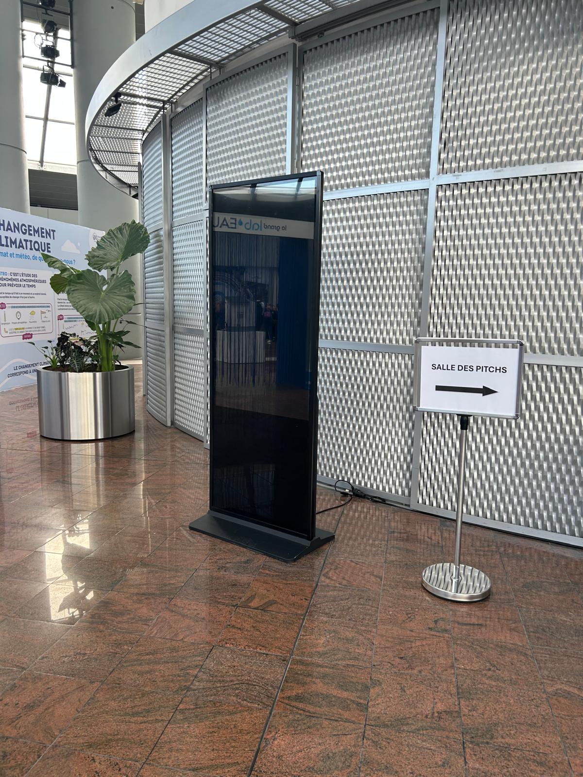Totem numérique noir vertical installé dans le hall d'un centre de congrès à côté d'un panneau directionnel