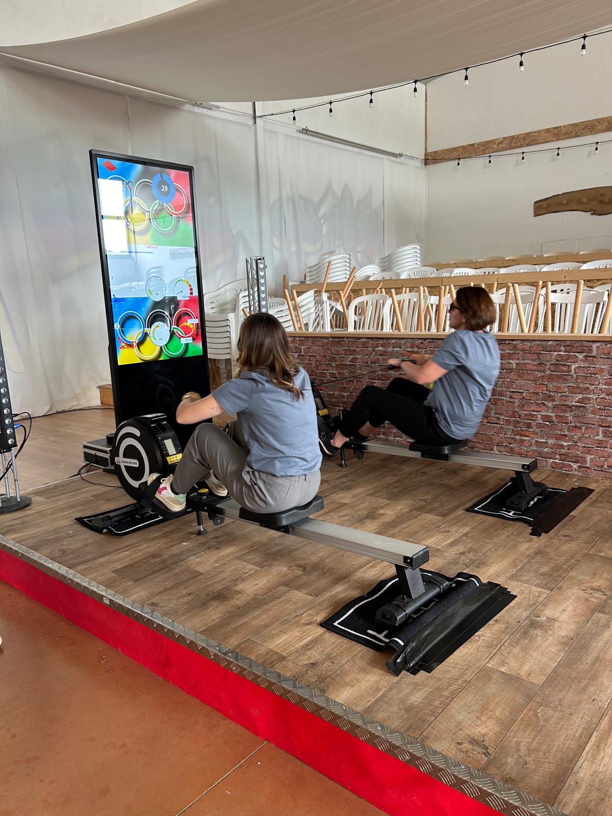 Deux participants s'affrontent sur des rameurs indoor face à un totem numérique dans une salle événementielle